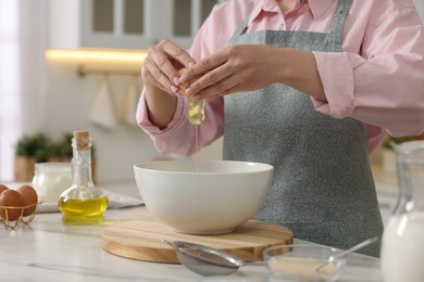 Making bread. Woman putting raw egg into bowl at white table in kitchen, closeup