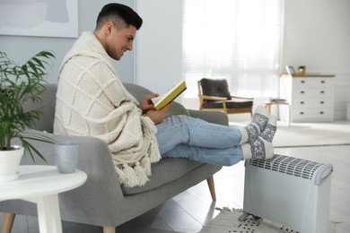 Photo of Happy man with book warming feet on electric heater at home