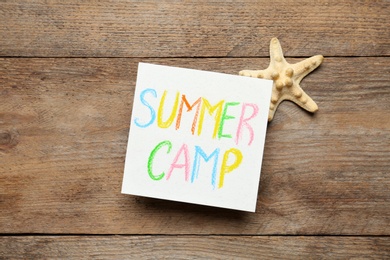 Card with text SUMMER CAMP and starfish on wooden table, flat lay