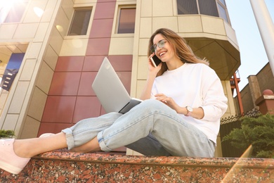 Image of Happy young woman talking on phone while using laptop outdoors, low angle view 