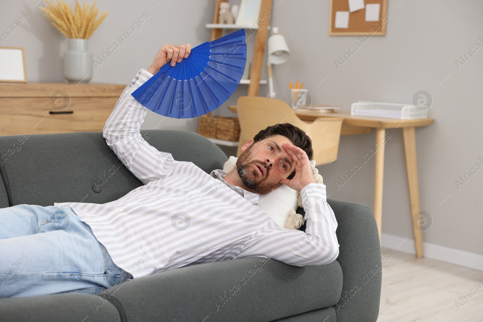 Photo of Bearded man waving blue hand fan to cool himself on sofa at home