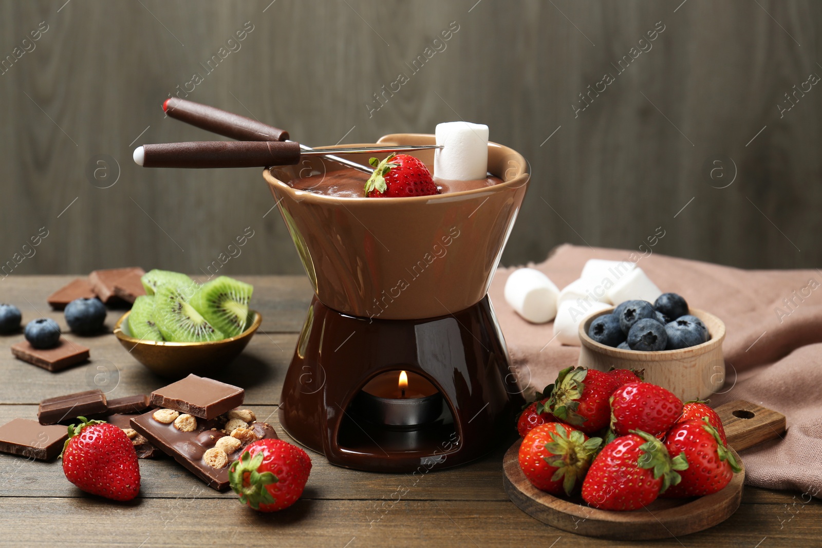 Photo of Fondue pot with melted chocolate, different fresh berries, kiwi, marshmallows and forks on wooden table