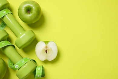 Photo of Fresh apples, measuring tape and dumbbells on green background, flat lay. Space for text