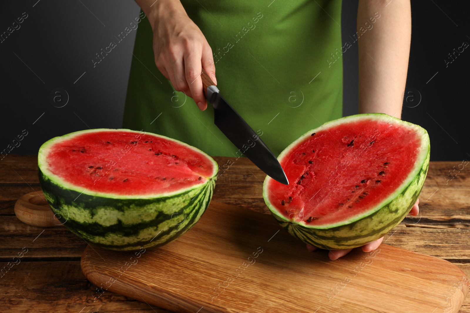 Photo of Woman cutting delicious watermelon at wooden table against dark grey background, closeup