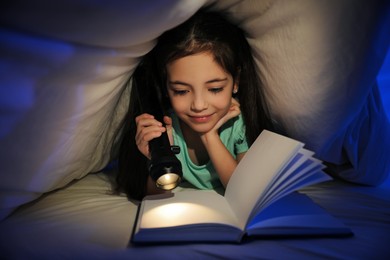 Photo of Little girl reading fairy tale under blanket in dark bedroom