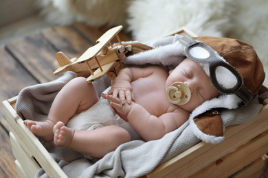 Cute newborn baby wearing aviator hat with toy sleeping in wooden crate