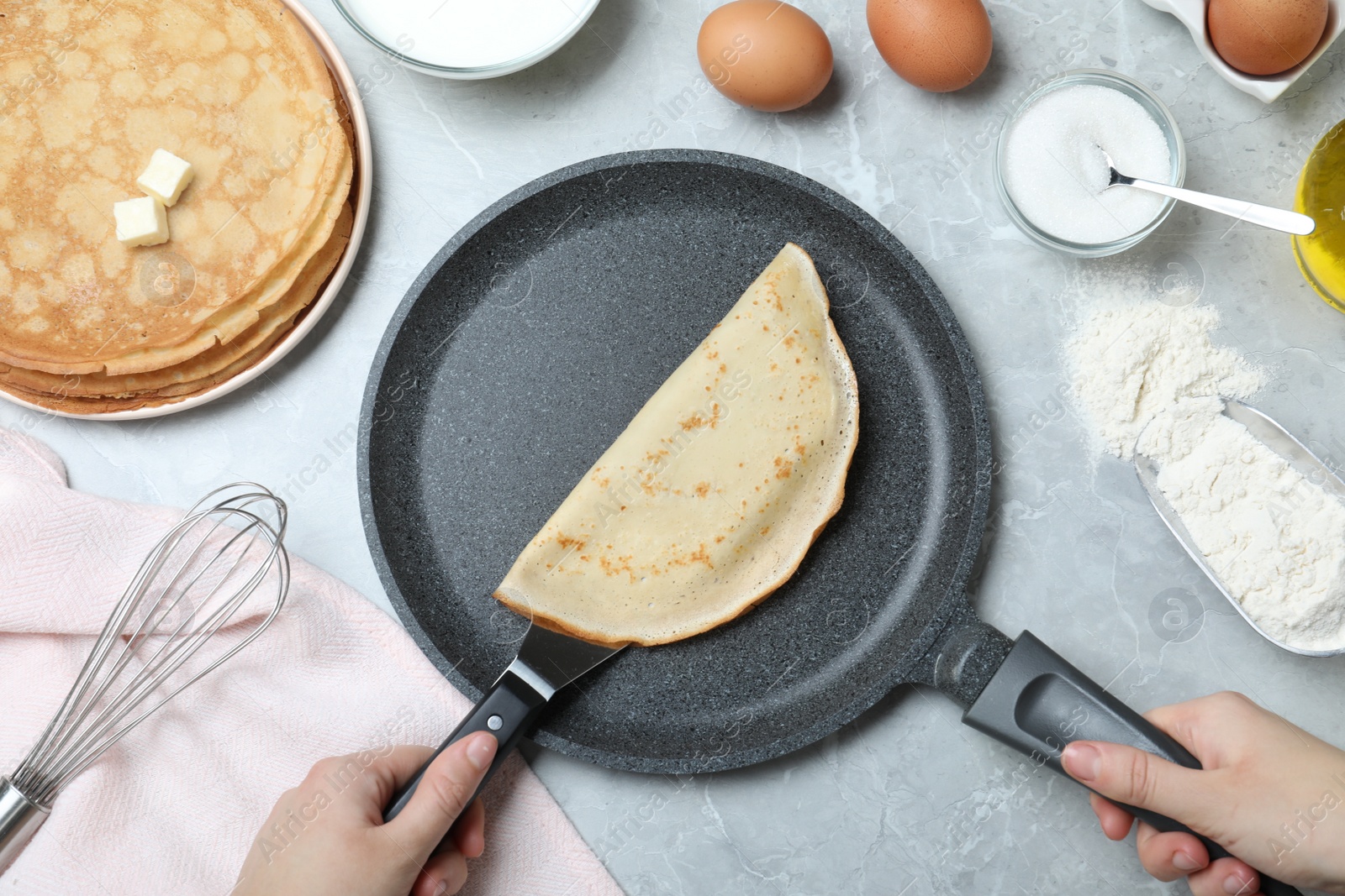 Photo of Woman holding frying pan with thin pancake at grey marble table, top view