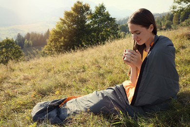 Young woman with drink in sleeping bag surrounded by beautiful nature