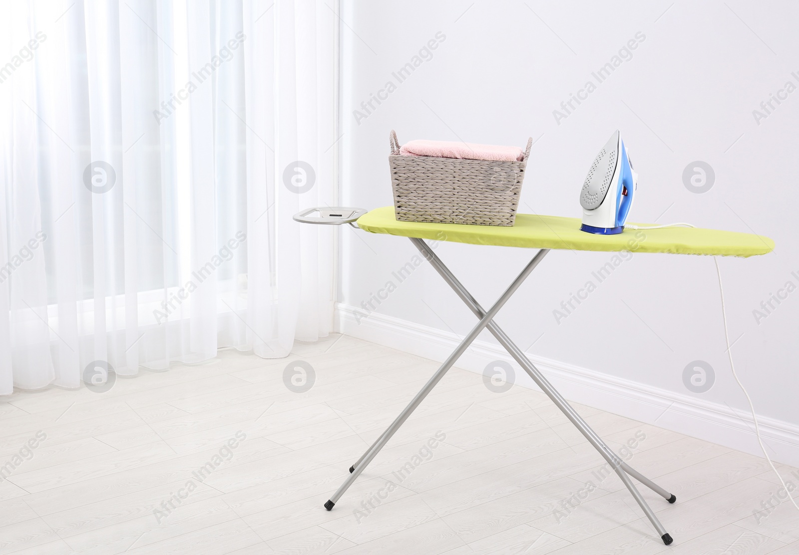 Photo of Board with modern iron and laundry basket in room