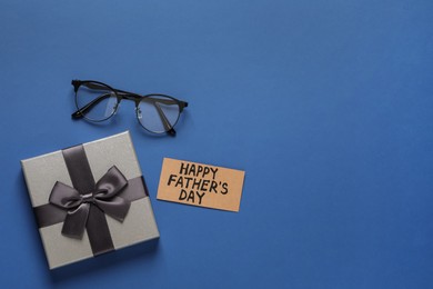 Photo of Card with phrase HAPPY FATHER'S DAY, eyeglasses and gift box on blue background, flat lay. Space for text