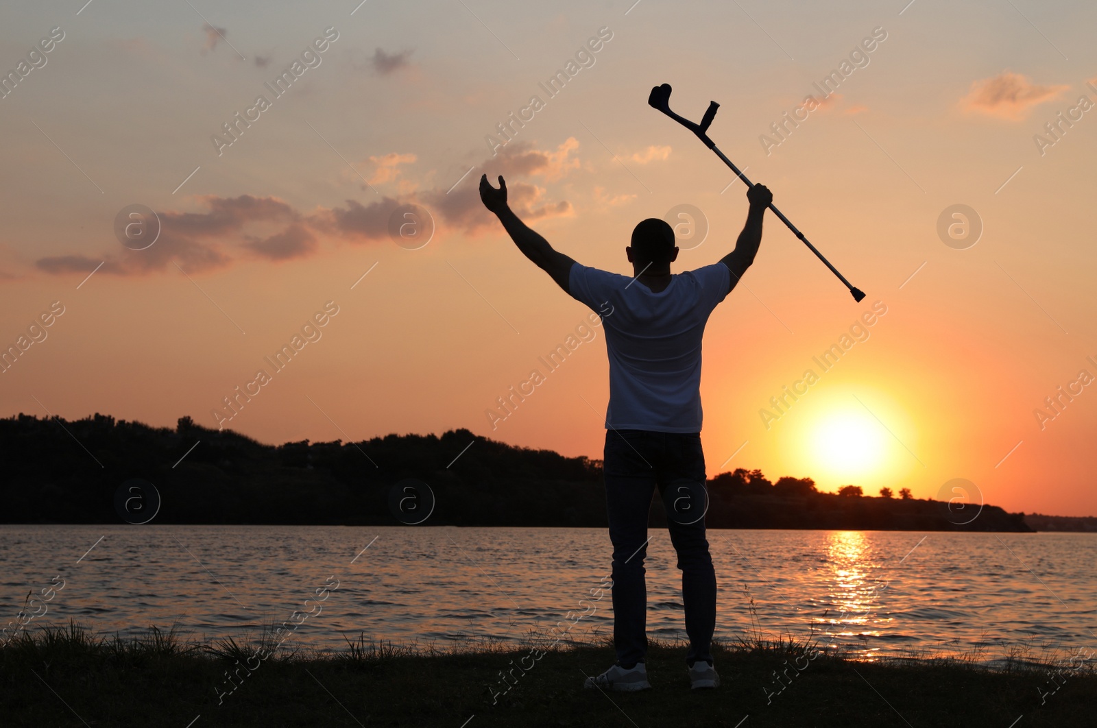 Photo of Man raising elbow crutch up to sky near river at sunset, back view. Healing miracle