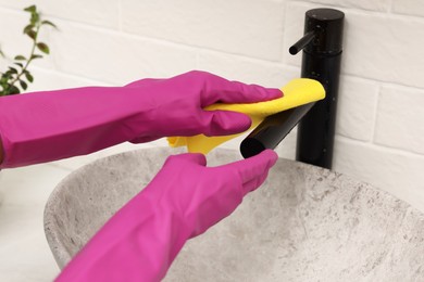 Photo of Woman in gloves cleaning faucet of bathroom sink with rag, closeup