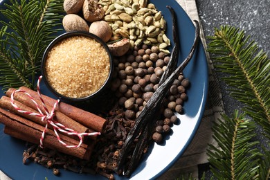 Plate with different aromatic spices and fir branches on grey textured table, top view