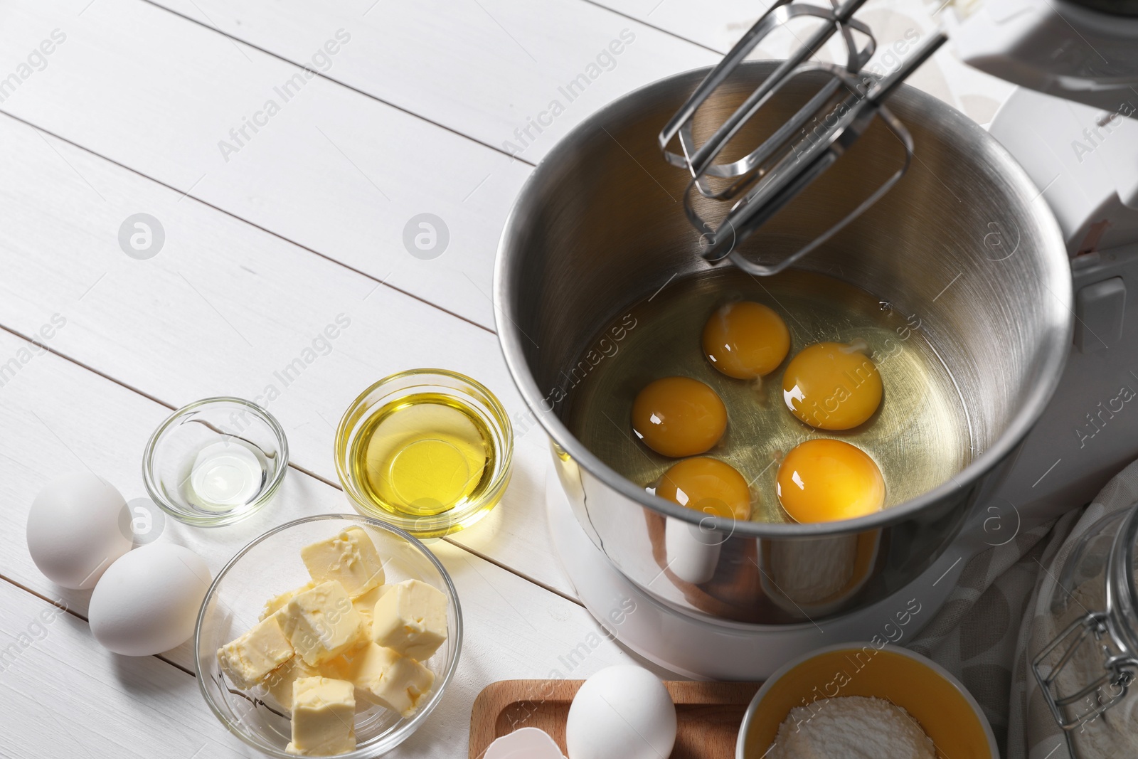 Photo of Making dough. Raw eggs in bowl of stand mixer and ingredients on white wooden table