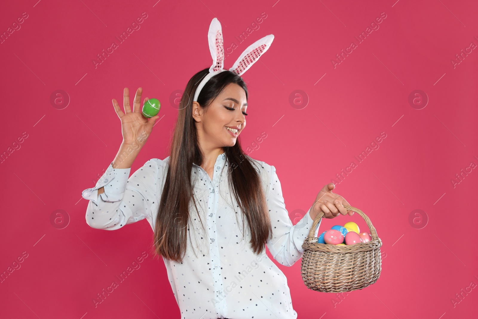 Photo of Beautiful woman in bunny ears headband with basket of Easter eggs on color background