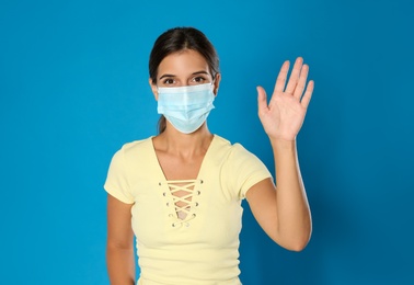 Photo of Woman in protective mask showing hello gesture on blue background. Keeping social distance during coronavirus pandemic