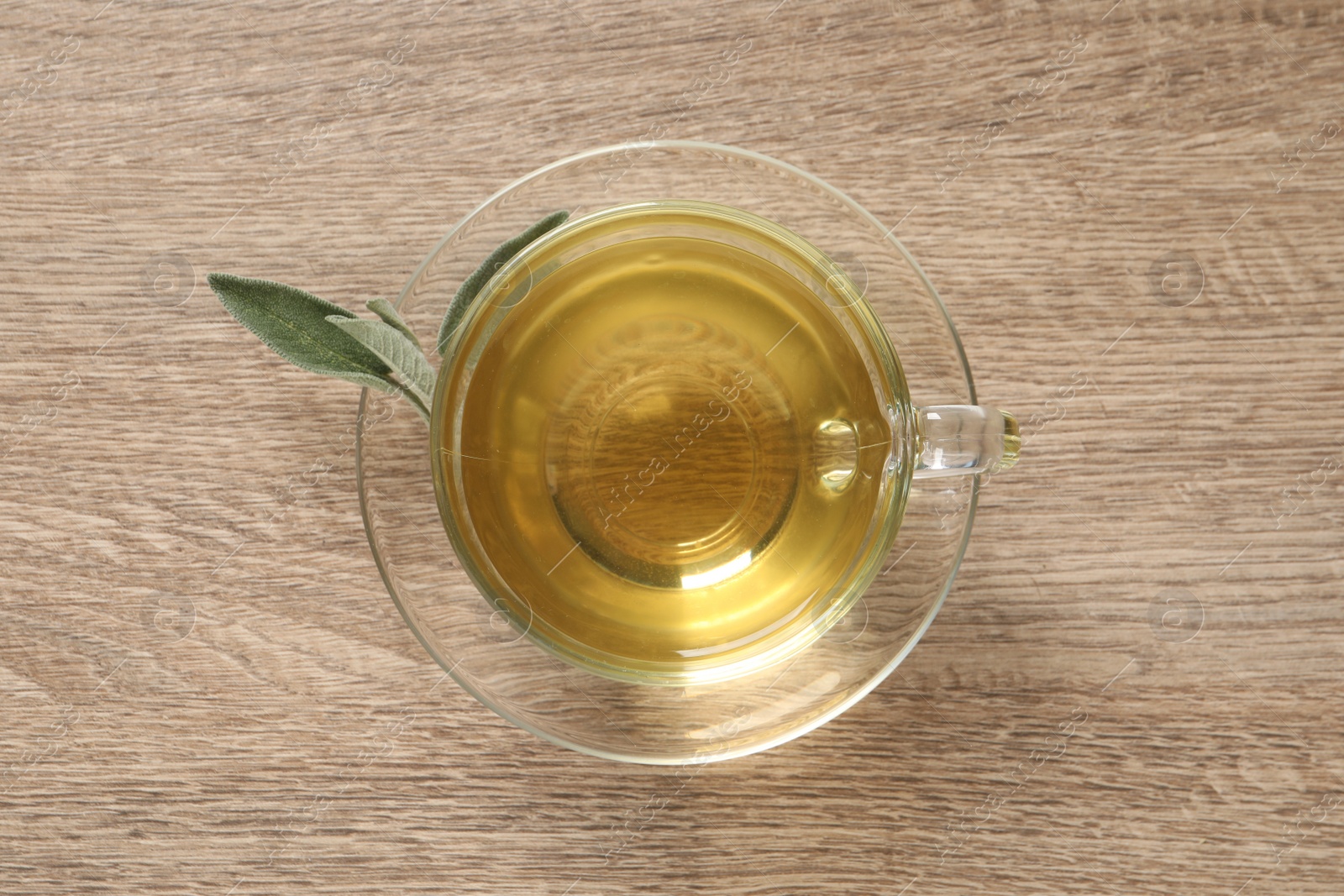 Photo of Cup of aromatic sage tea and fresh leaves on wooden table, top view