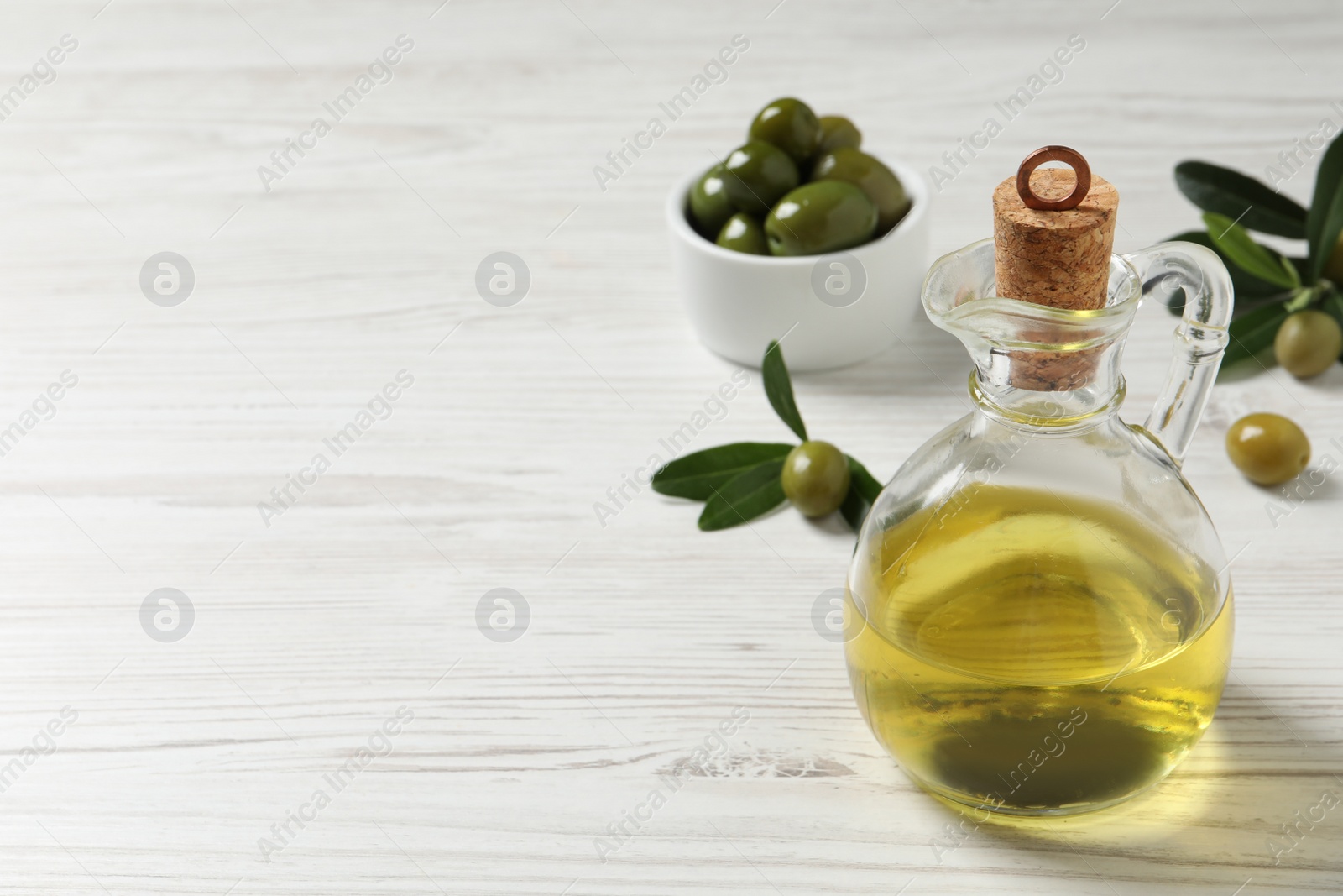 Photo of Glass jug of oil, ripe olives and green leaves on white wooden table. Space for text