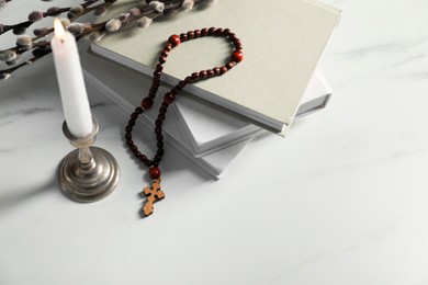 Books, burning candle, willow branches and rosary beads on white marble table, space for text