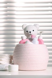 Wicker basket with baby cosmetic products and knitted toy bear on white table