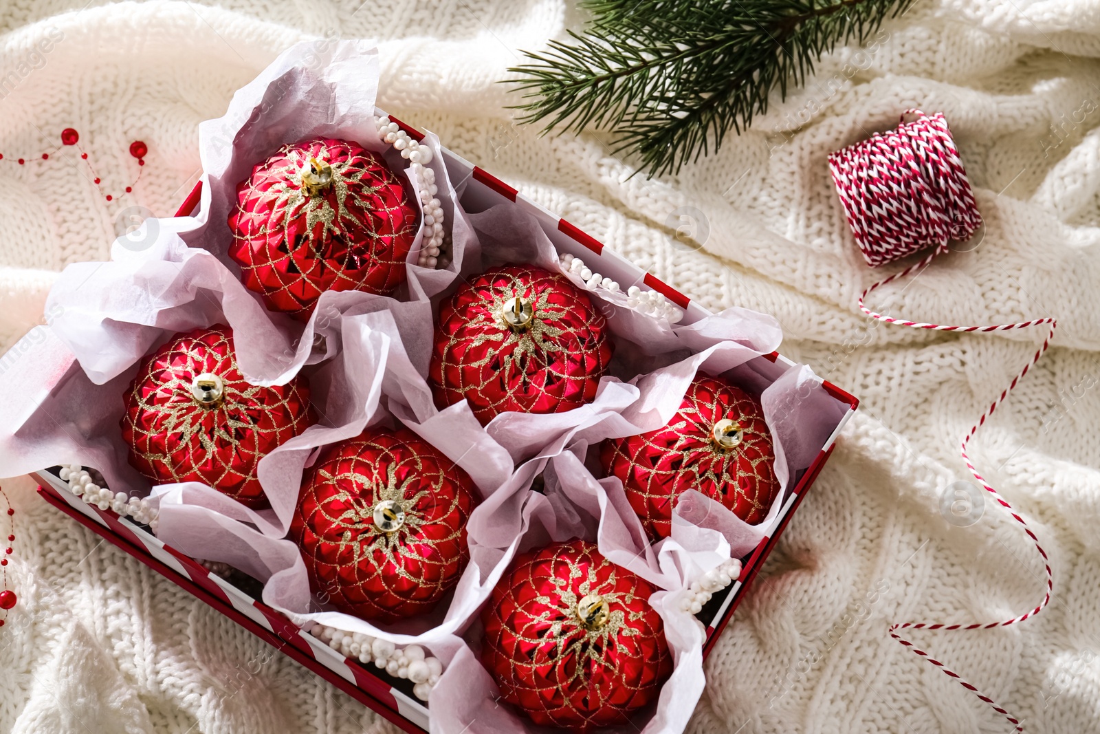 Photo of Beautiful red Christmas baubles in box on knitted plaid, flat lay