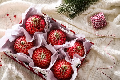 Photo of Beautiful red Christmas baubles in box on knitted plaid, flat lay
