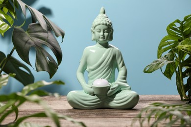 Buddhism religion. Decorative Buddha statue with burning candle on wooden table and houseplants against light blue wall, selective focus