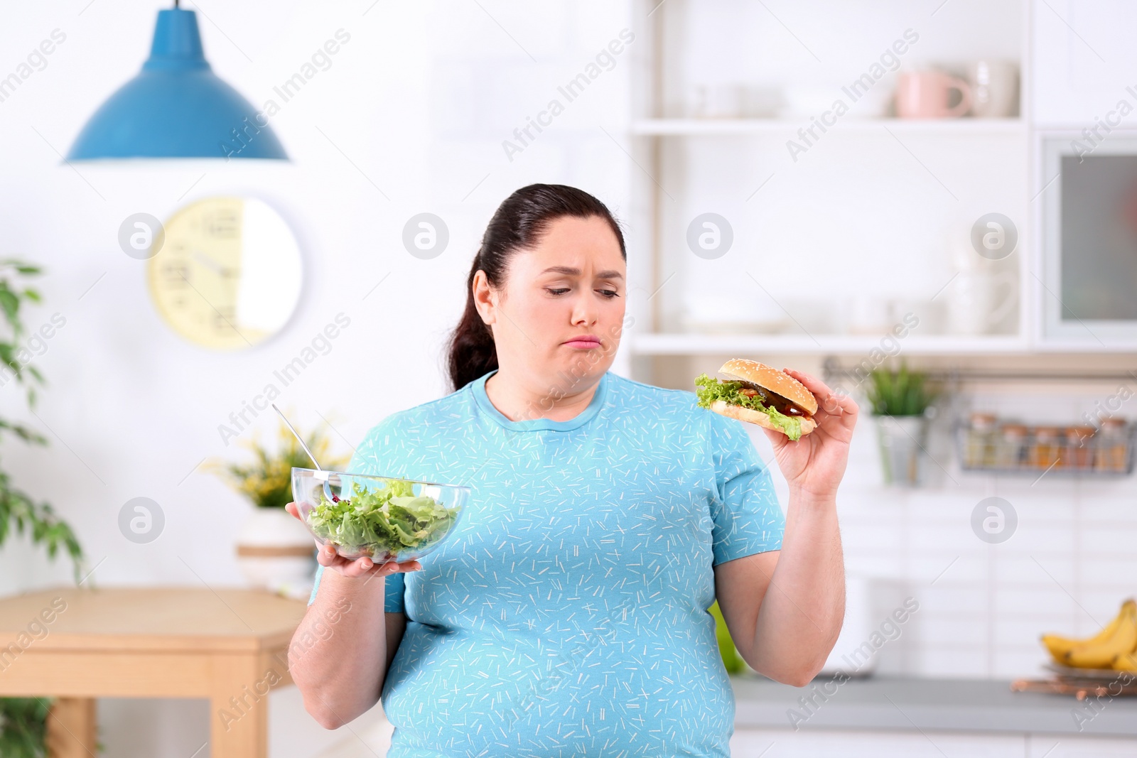 Photo of Sad overweight woman with salad and burger in kitchen. Healthy diet