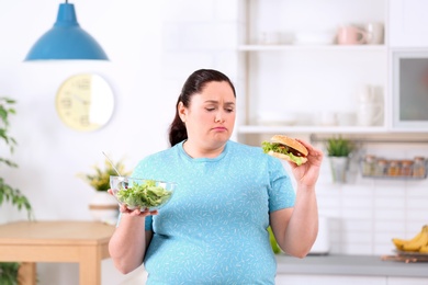 Photo of Sad overweight woman with salad and burger in kitchen. Healthy diet