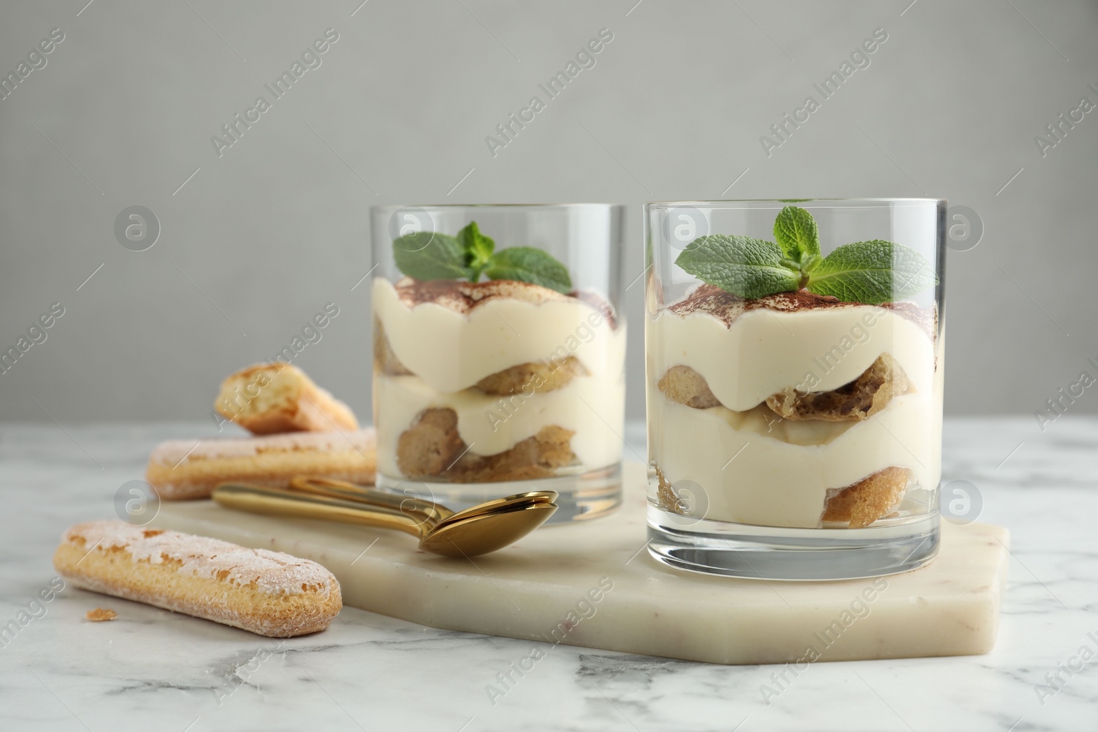 Photo of Delicious tiramisu in glasses, mint leaves, cookies and spoons on white marble table