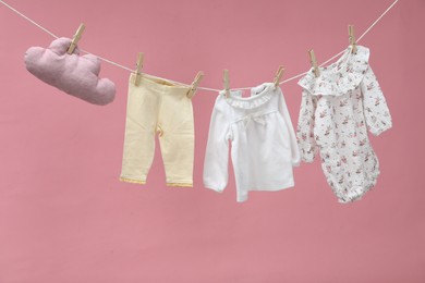 Different baby clothes and cloud shaped pillow drying on laundry line against pink background