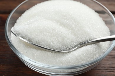 Photo of Granulated sugar in bowl and spoon on wooden table, closeup