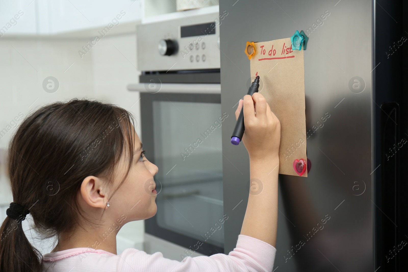 Photo of Little girl writing to do list on fridge in kitchen