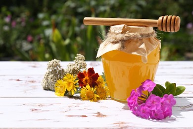 Photo of Glass jar of honey, dipper and different flowers on white wooden table in garden. Space for text