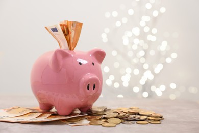 Photo of Piggy bank with euro banknotes and coins on grey table against blurred lights, space for text