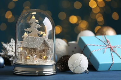 Photo of Beautiful snow globe, gift box and Christmas decor on blue table against blurred festive lights
