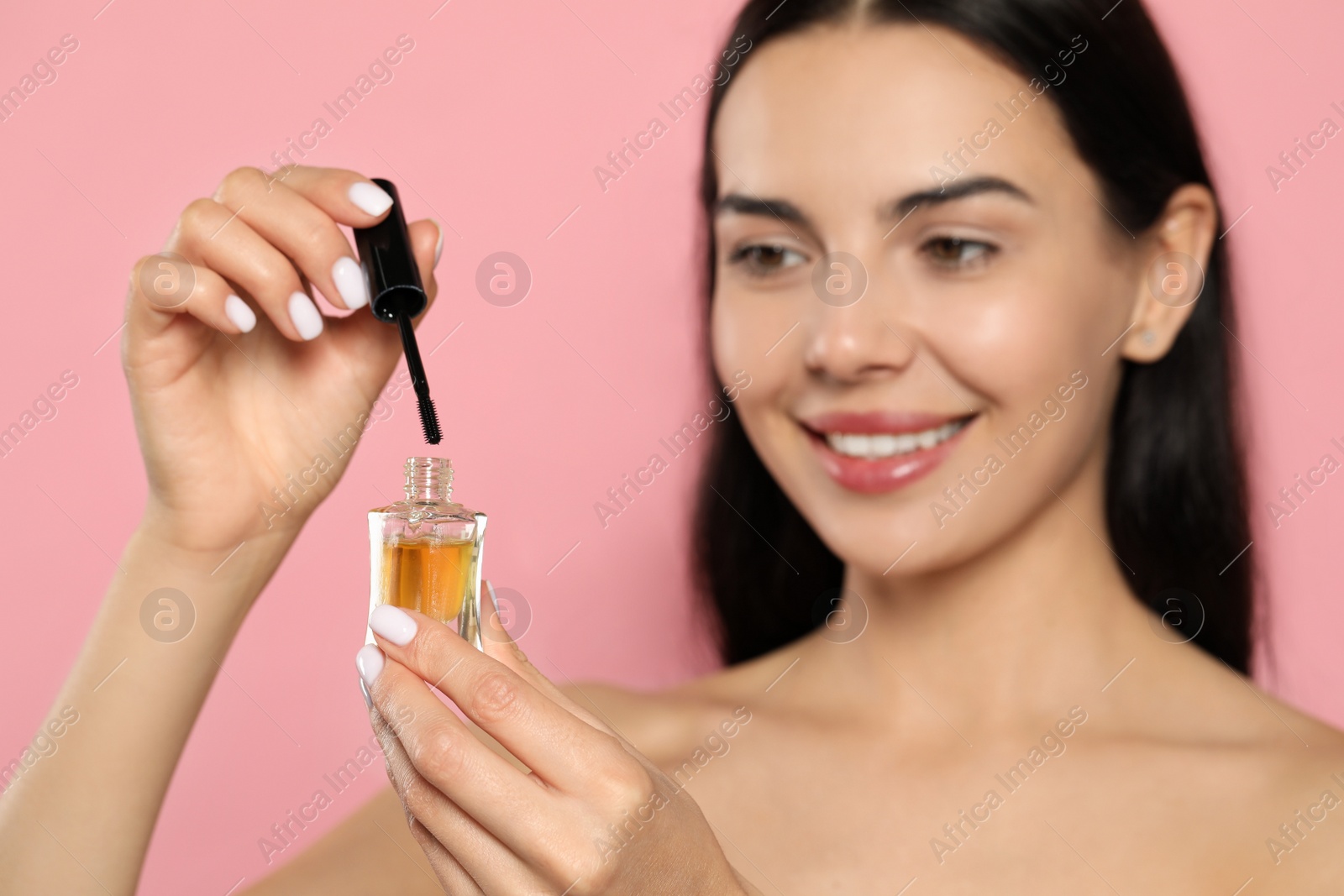 Photo of Beautiful young woman with eyelash oil against pink background, focus on bottle