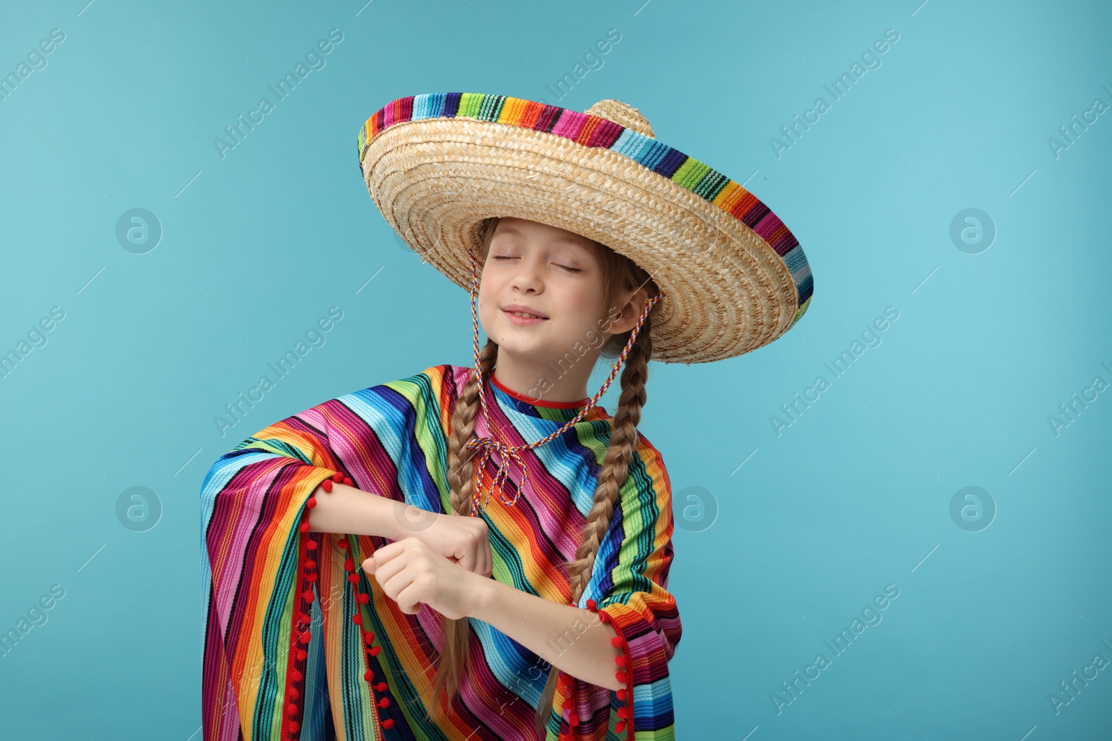 Photo of Cute girl in Mexican sombrero hat and poncho dancing on light blue background