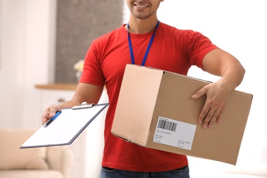 Photo of Young courier holding parcel and clipboard indoors, closeup. Delivery service