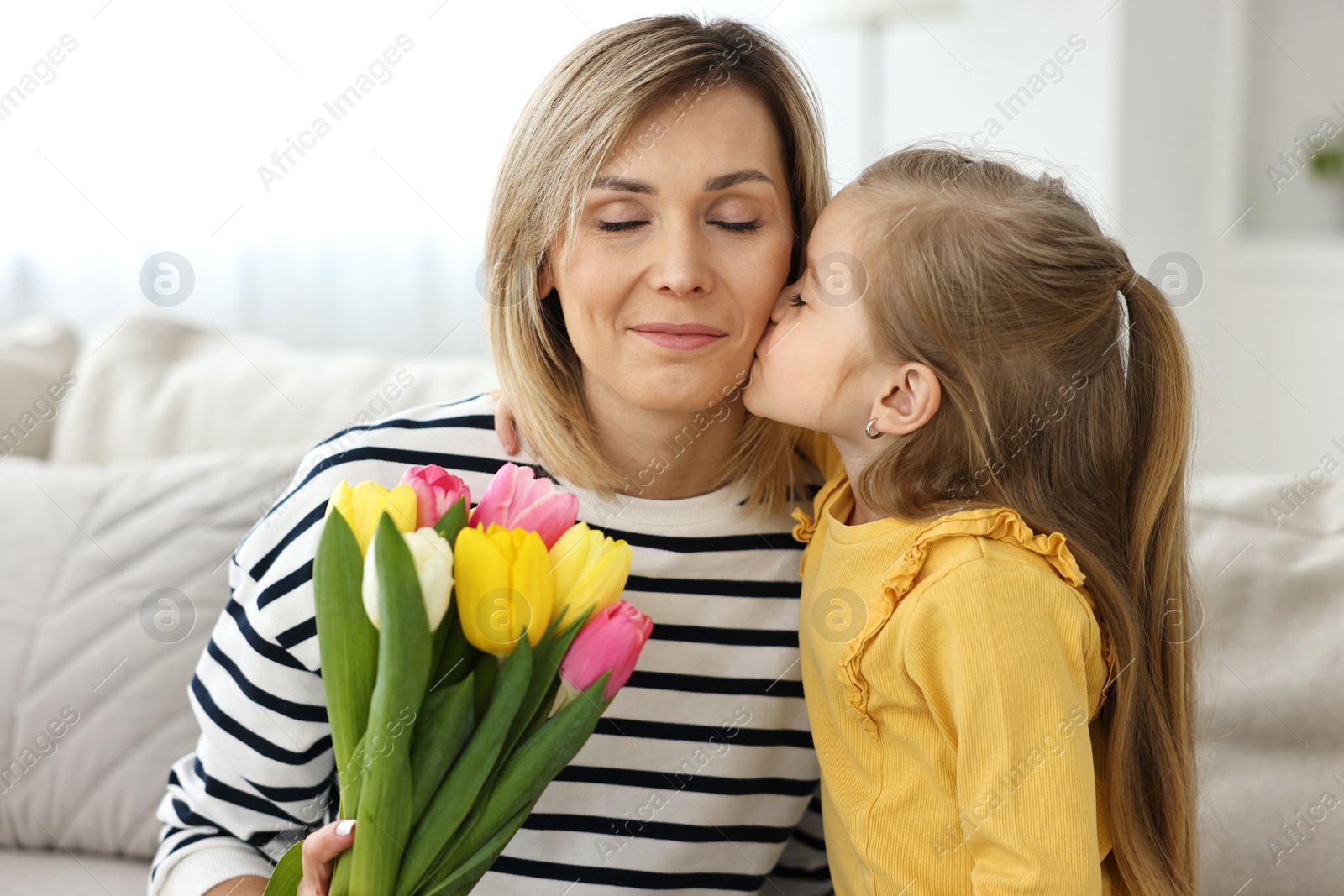 Photo of Little daughter kissing and congratulating her mom with Mother`s Day at home. Woman holding bouquet of beautiful tulips