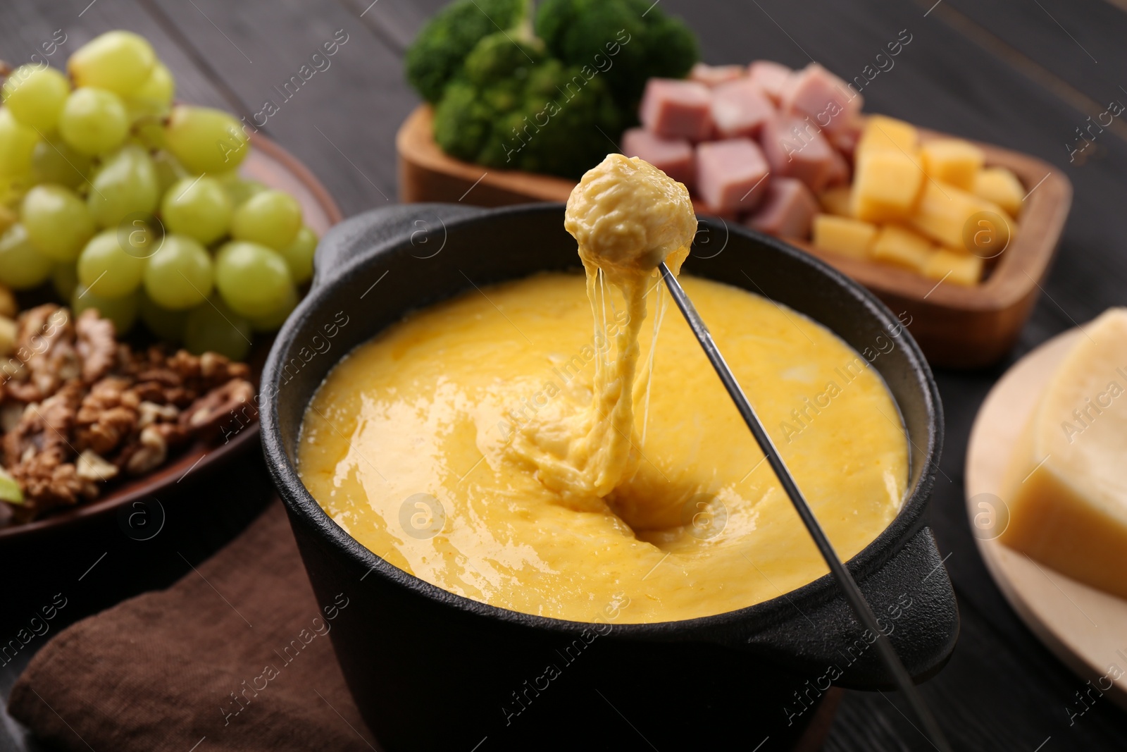 Photo of Dipping piece of ham into fondue pot with melted cheese on table, closeup