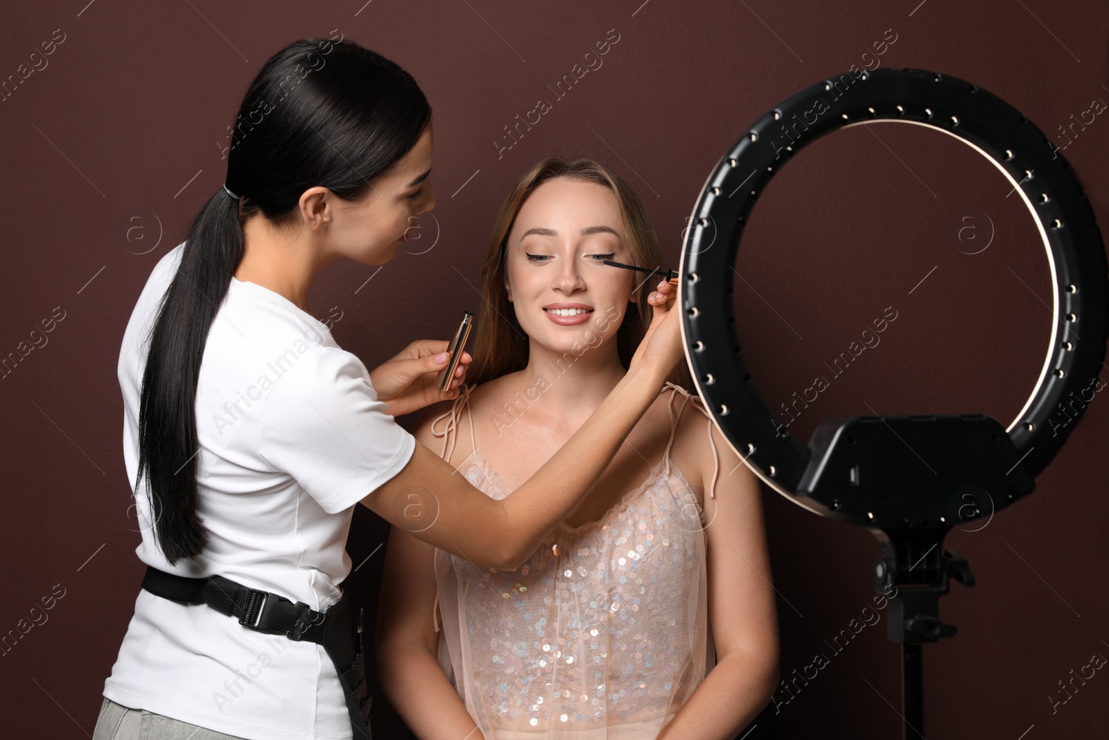 Photo of Professional makeup artist working with beautiful young woman against brown background. Using ring lamp