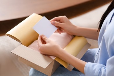 Woman holding blank greeting card near package with gift indoors, closeup