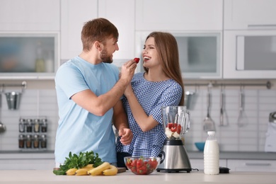 Photo of Young couple eating strawberries while preparing delicious milk shake in kitchen