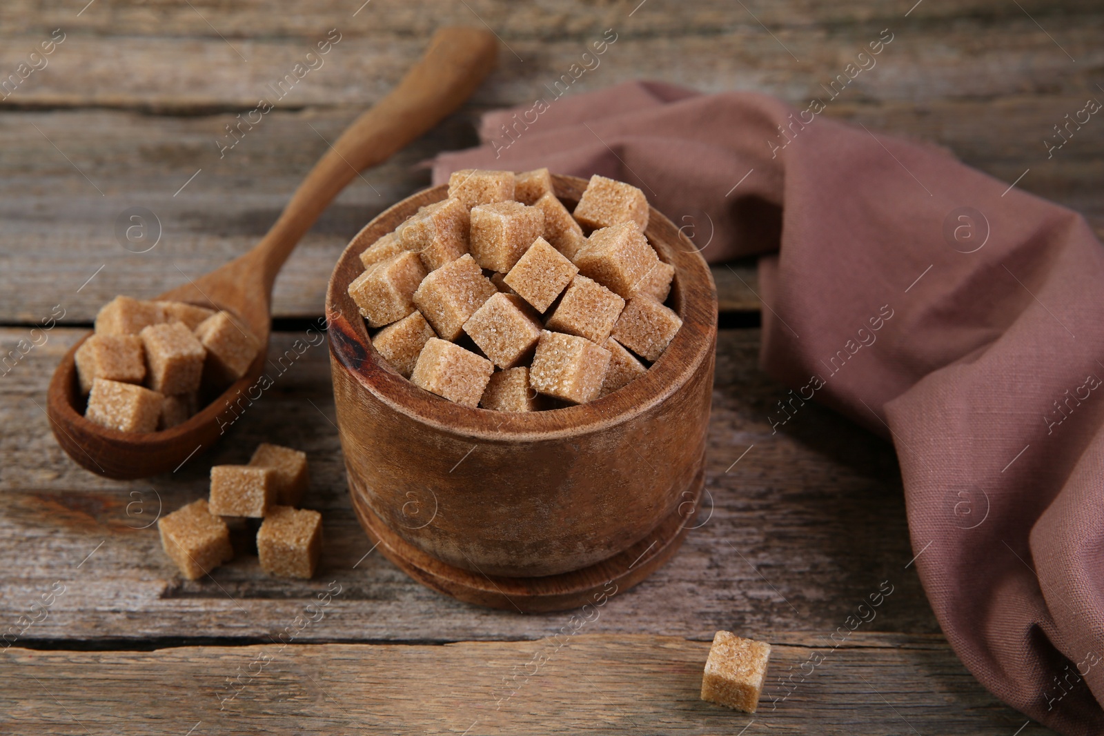 Photo of Brown sugar cubes in bowl and spoon on wooden table, closeup