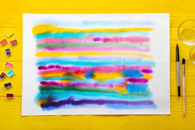Photo of Bright abstract watercolor painting, paints, empty bowls and brushes on yellow wooden table, flat lay