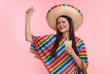 Photo of Young woman in Mexican sombrero hat and poncho holding cocktail while dancing on pink background