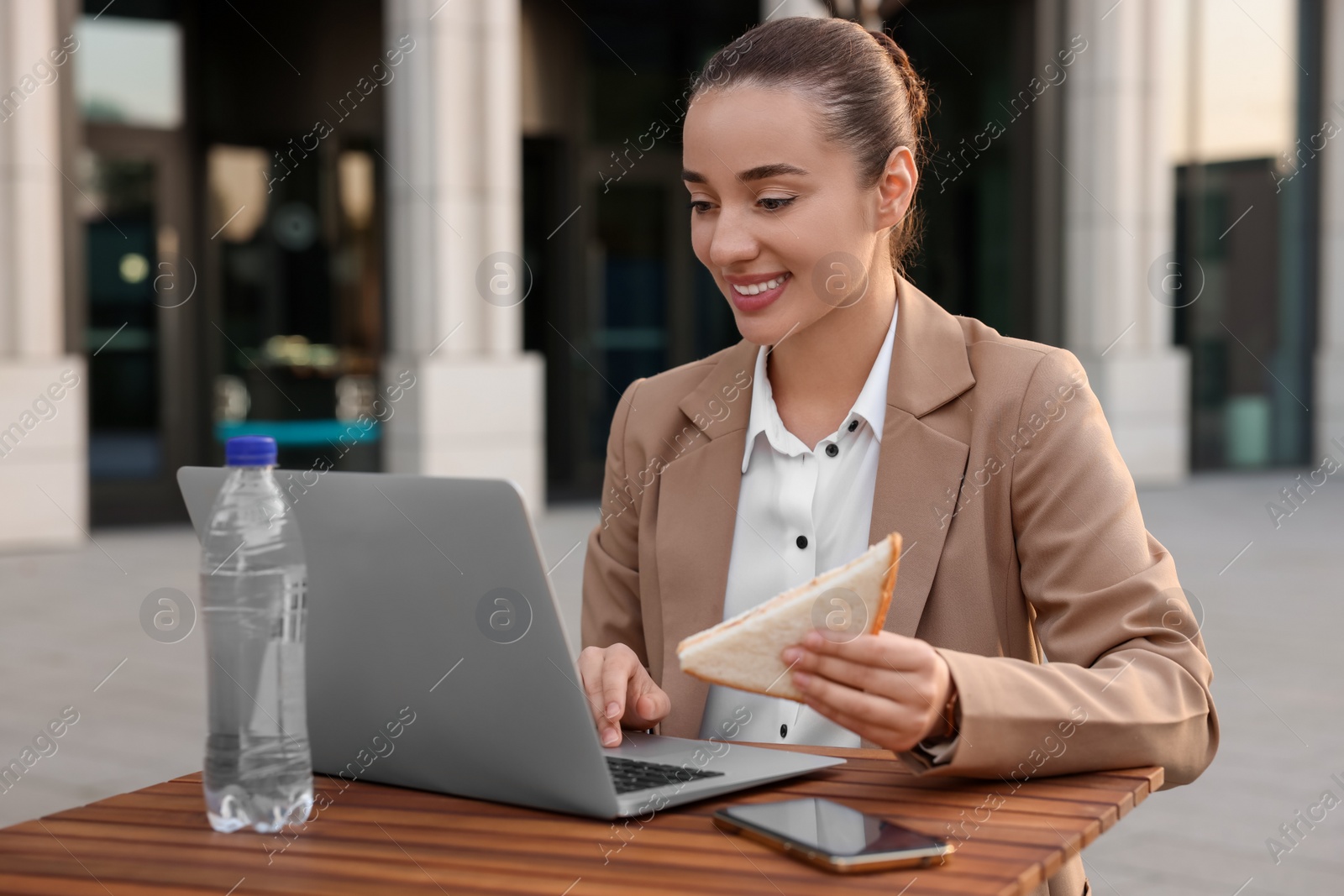 Photo of Happy businesswoman with sandwich using laptop during lunch at wooden table outdoors