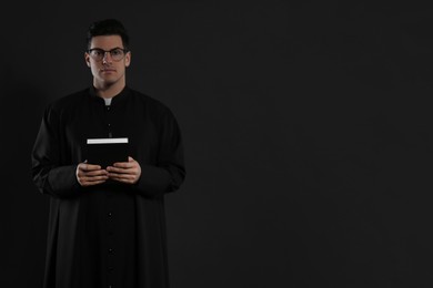 Priest with Bible on black background. Space for text