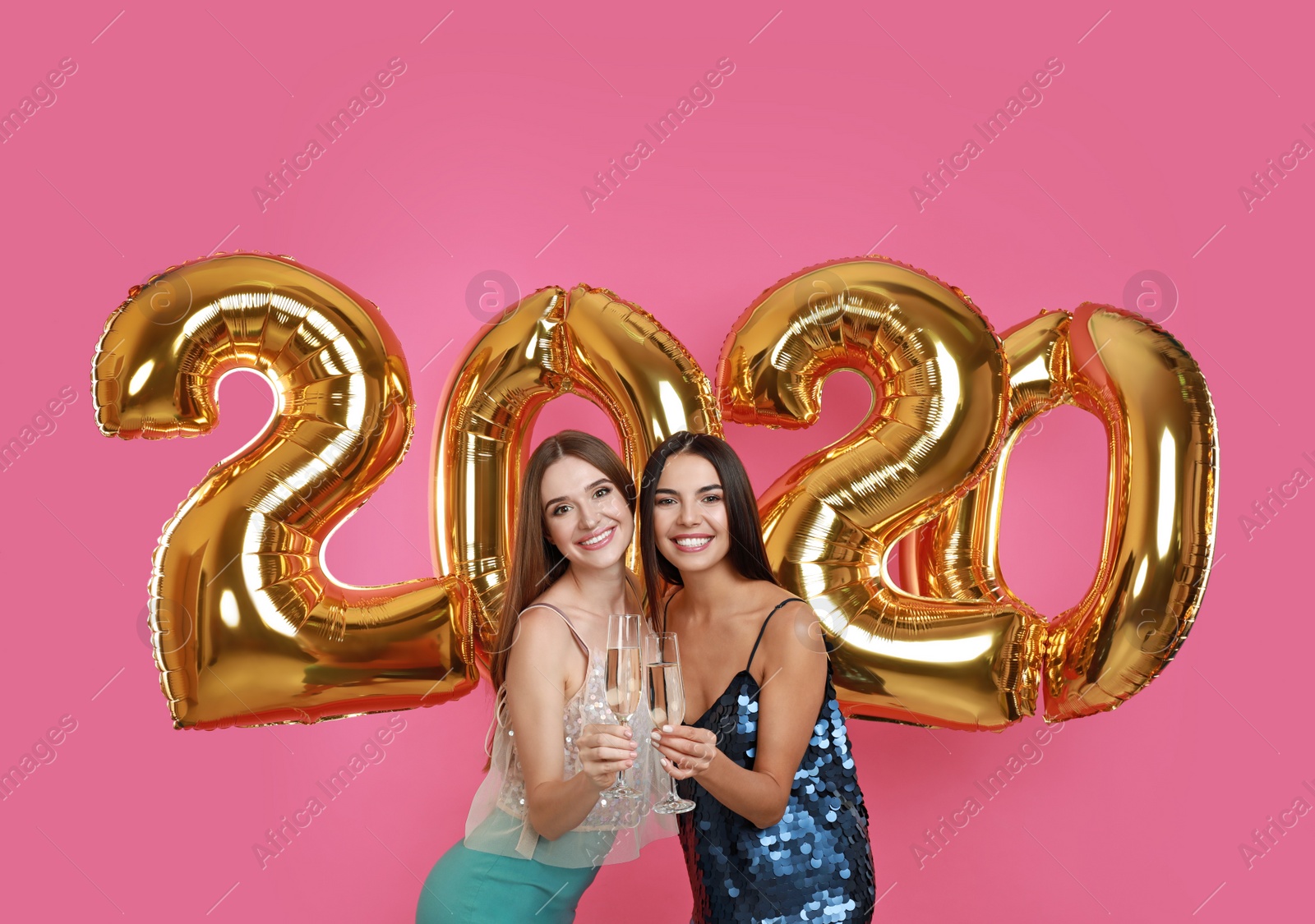 Photo of Happy young women with glasses of champagne near golden 2020 balloons on pink background. New Year celebration